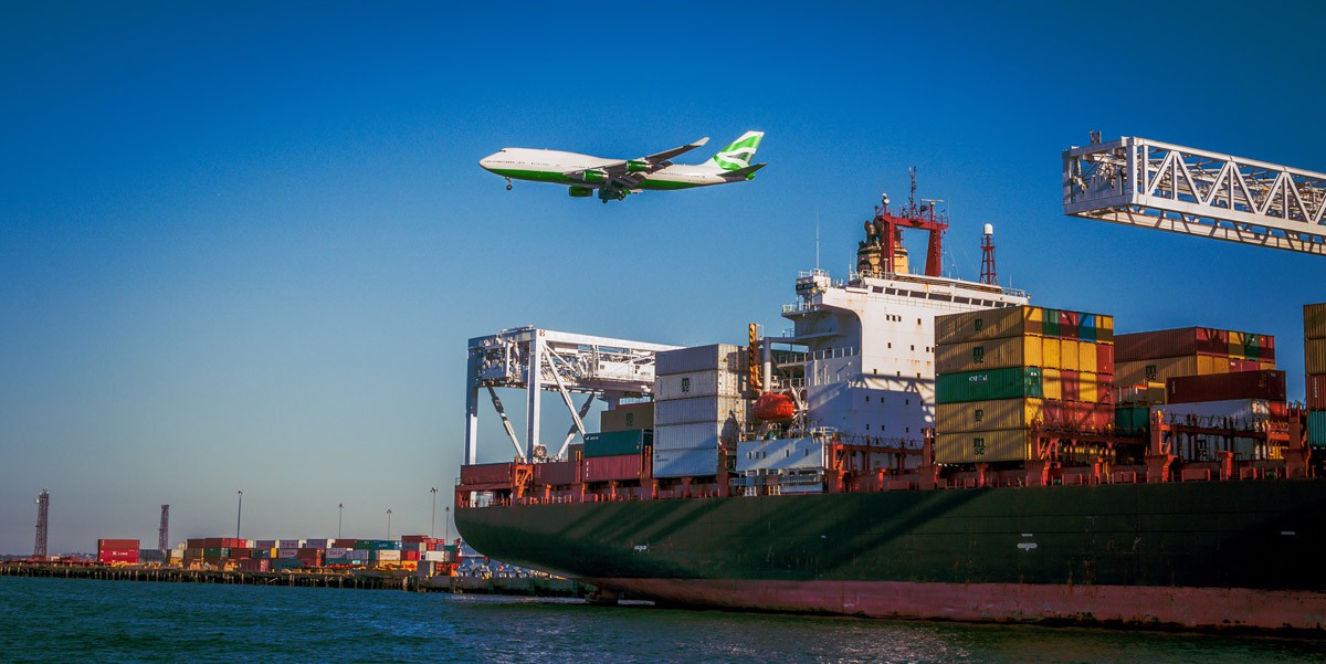 A plane flies over a ship carrying large shipping cargo containers.