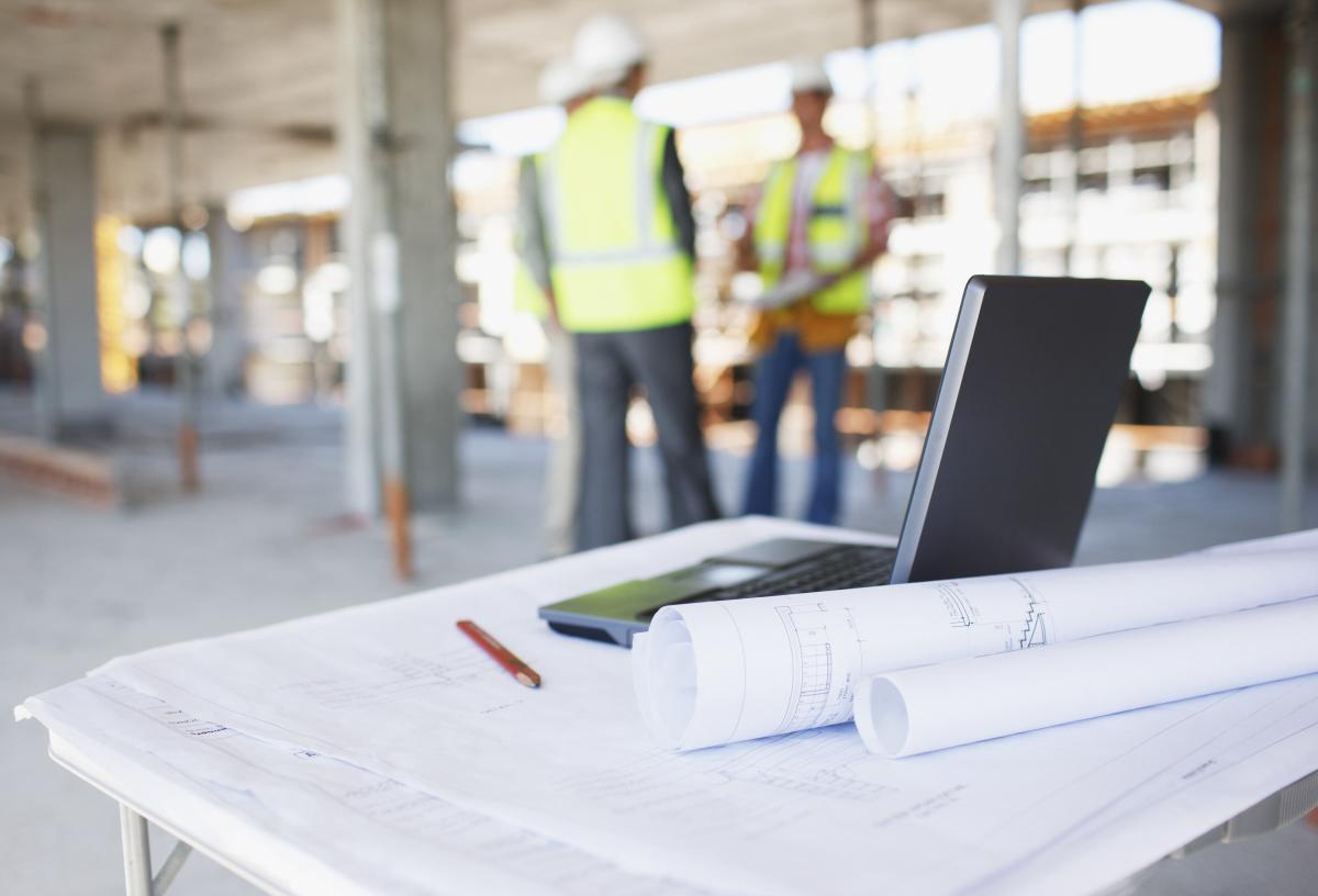 Contractors stand at a construction site, talking. In the foreground, there are blueprints and a laptop on a table.