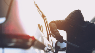 A hooded thief using a crowbar to break into a car.